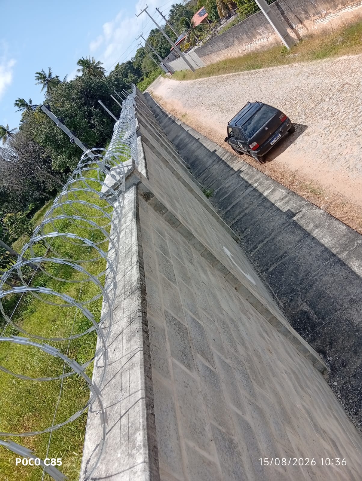 Vista lateral de muro extenso com concertina instalada, protegendo perímetro ao longo de via de acesso